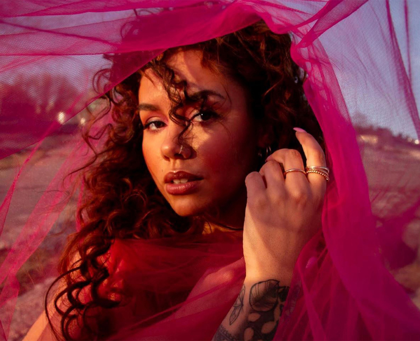 Woman with lustrous curly hair draped in pink veil at sunset